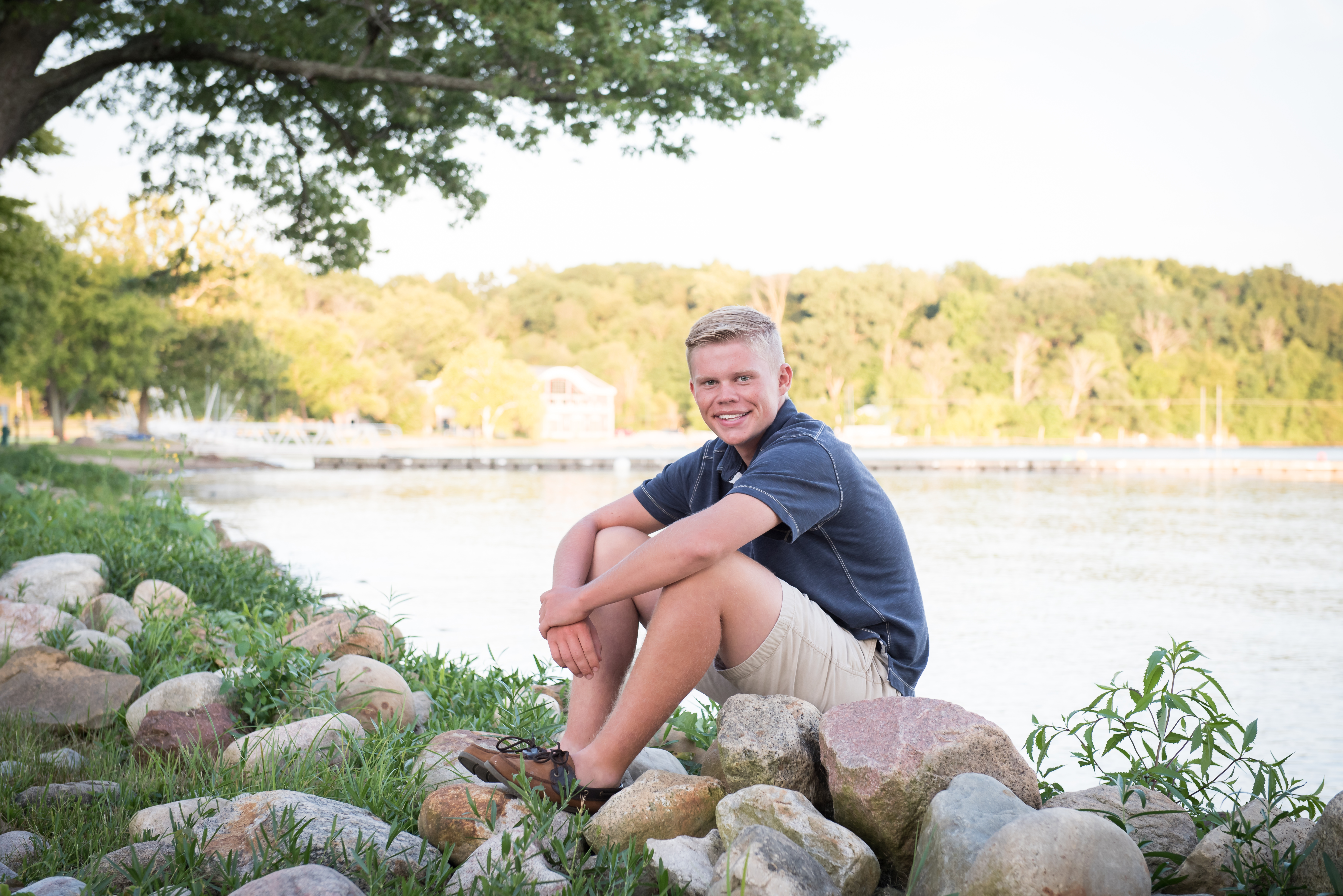 Senior Portrait at Culver Academies sitting on rocks by Lake Maxinkuckee for pictures in blue shirt and khaki pants. 