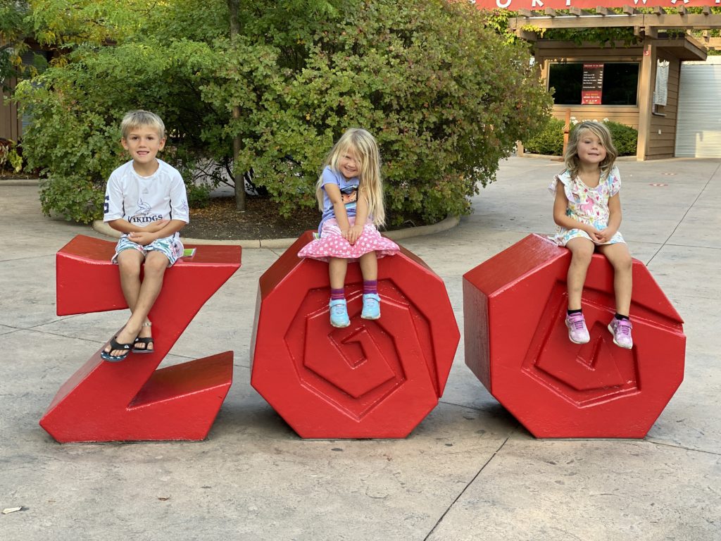Three little kids sitting on red zoo sign at the entrance to the Fort Wayne Zoo in Indiana. 