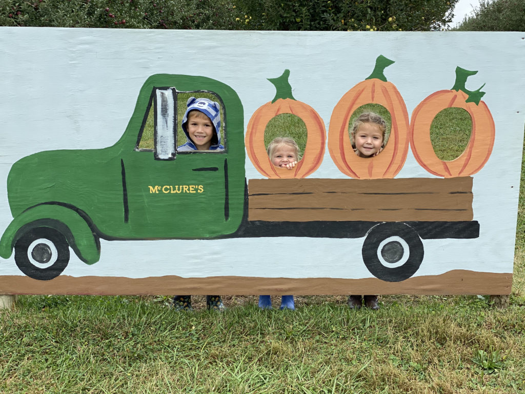 Three kids in McClure's apple orchard sign smiling in the play area. 