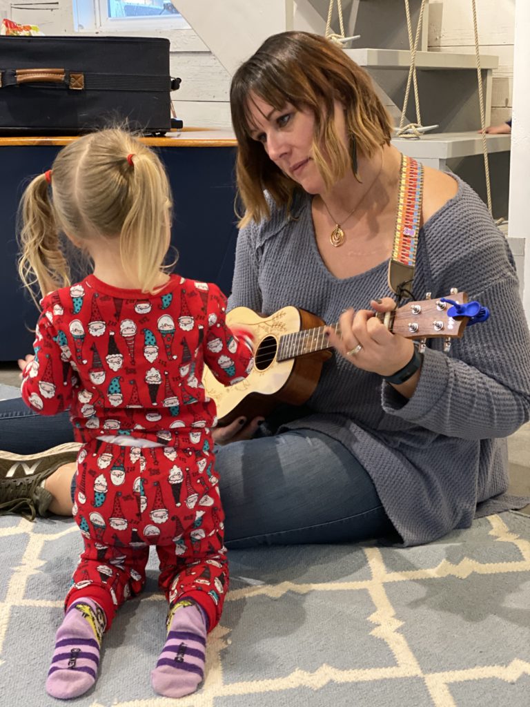 Little girl wearing Christmas pajamas in Music for Me Class with Wendelyn Daly strumming Ukelele Ms. Wendy holds, while sitting on the floor in the Always Images Studio location of the class. 