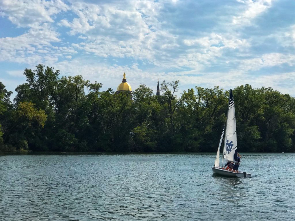Notre Dame sailboat on the campus lake with the Golden Dome in the background and blue skies with trees. 