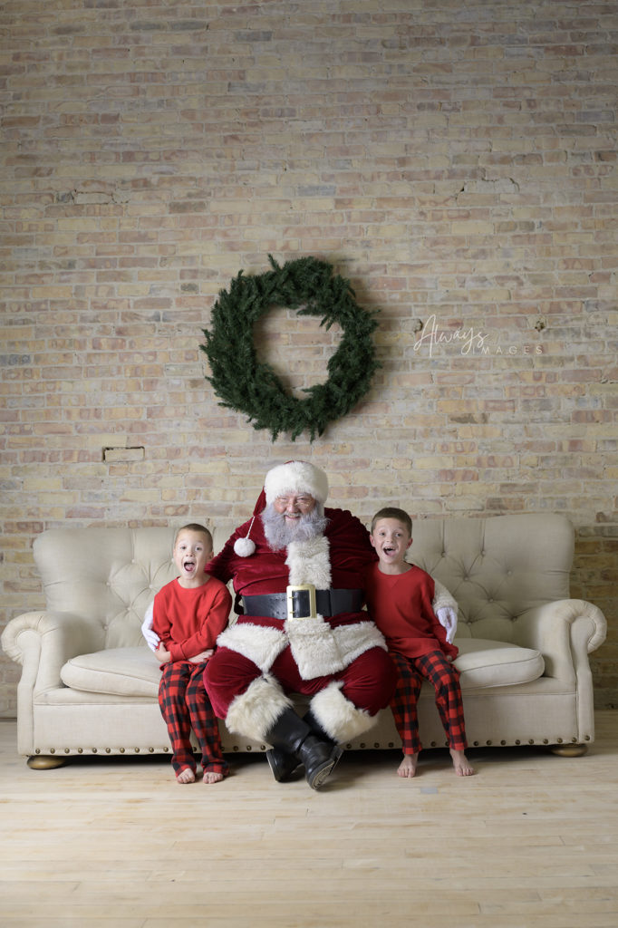 Kids in red pajamas with Santa and a wreath sitting on a couch with brick walls behind them. 