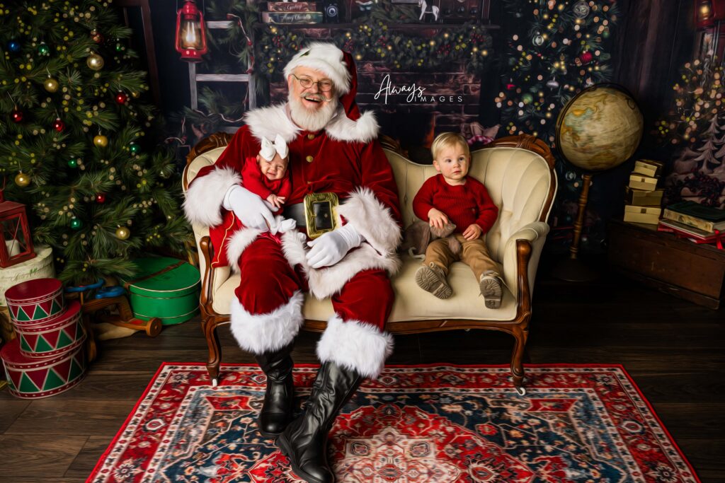 Baby sitting on Santa's lap with toddler boy next to Santa on an antique cream colored couch. 