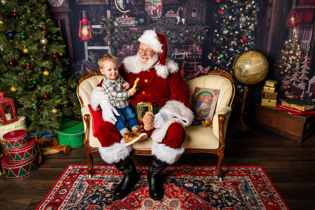 Toddler boy dressed in jeans and plaid shirt sitting on Santa's lap on a cream antique couch in Santa's living room set in a photography studio. 